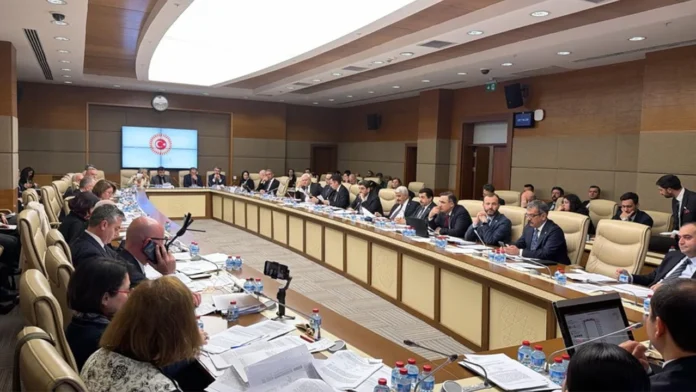 TBMM Komisyon salonunda, ön plandaki ahşap masada açık duran bir dizüstü bilgisayar ve yanında bir not kağıdı ile kalem. Bilgisayar ekranında oyun platformu arayüzü görünüyor. Arka planda, duvarda asılı büyük Türk bayrağı ve toplantı düzeninde yer alan kırmızı koltuklar, loş ve doğal bir ışıklandırmayla, ciddi bir toplantı sonrası atmosferini yansıtıyor.